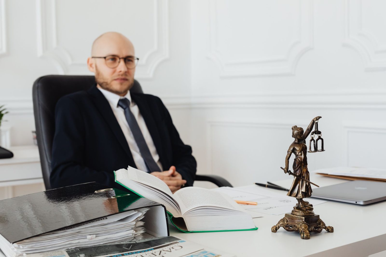Un abogado calvo sentado en un escritorio con documentos legales junto a una estatua de la Justicia.