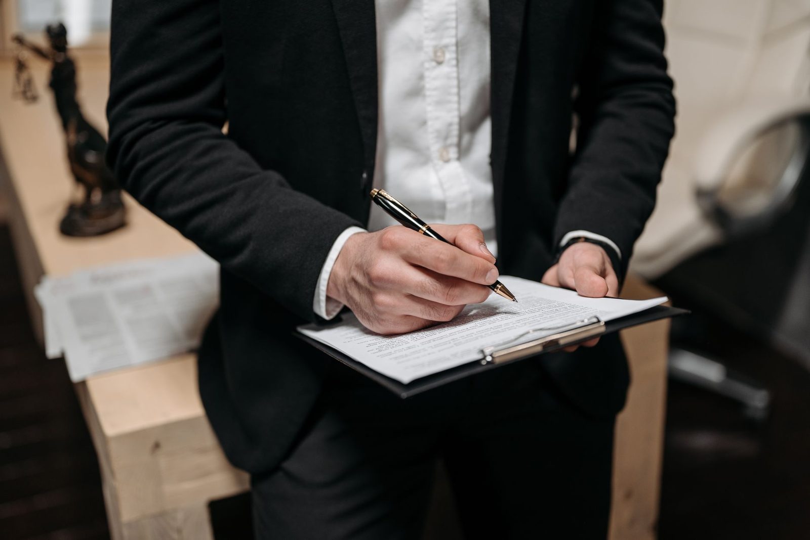 A business professional signs a contract on a clipboard at a desk in an office.