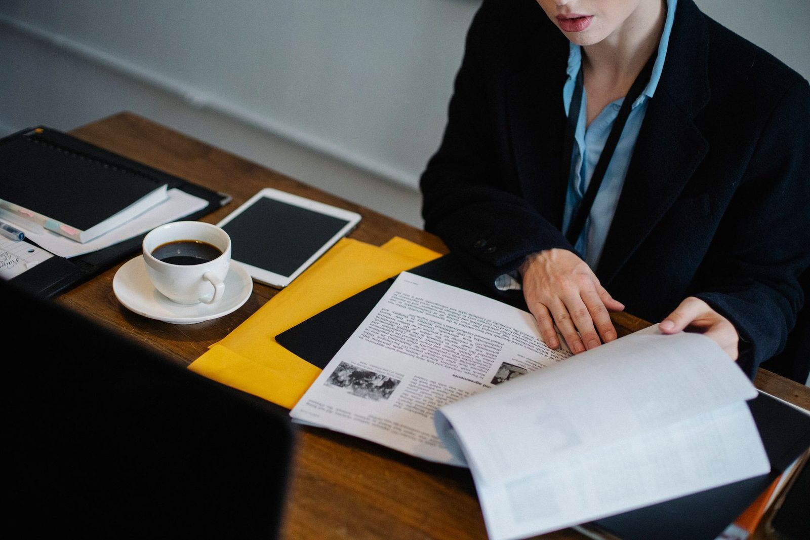 A businesswoman in formal attire reviews documents and a tablet at a desk with a cup of coffee.