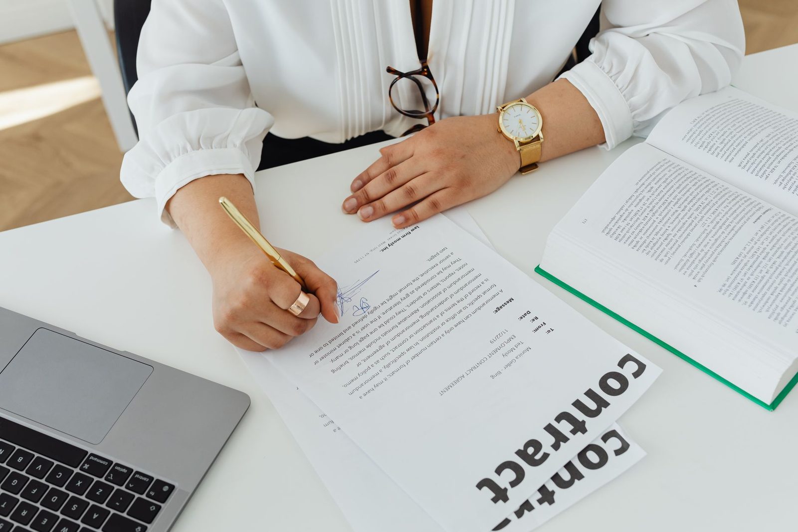 Una mujer de negocios firma un contrato en un escritorio junto a una laptop y un libro.