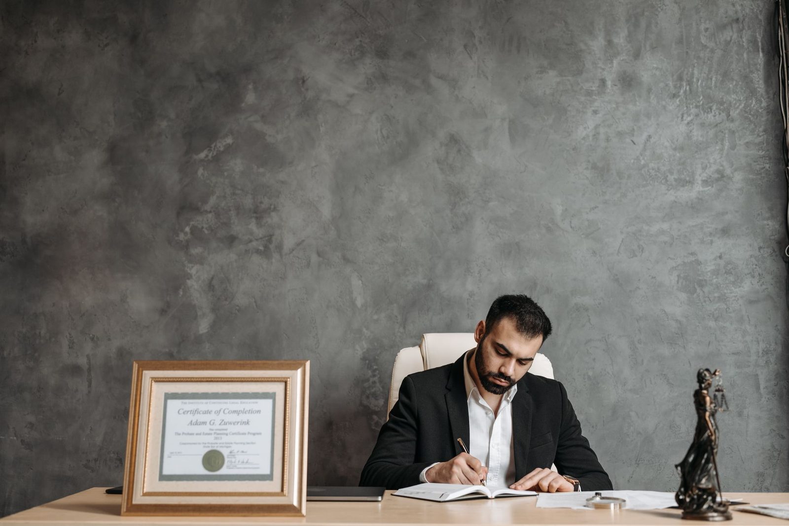Una empresaria escribiendo en un escritorio junto a un certificado enmarcado y una estatua de la justicia.
