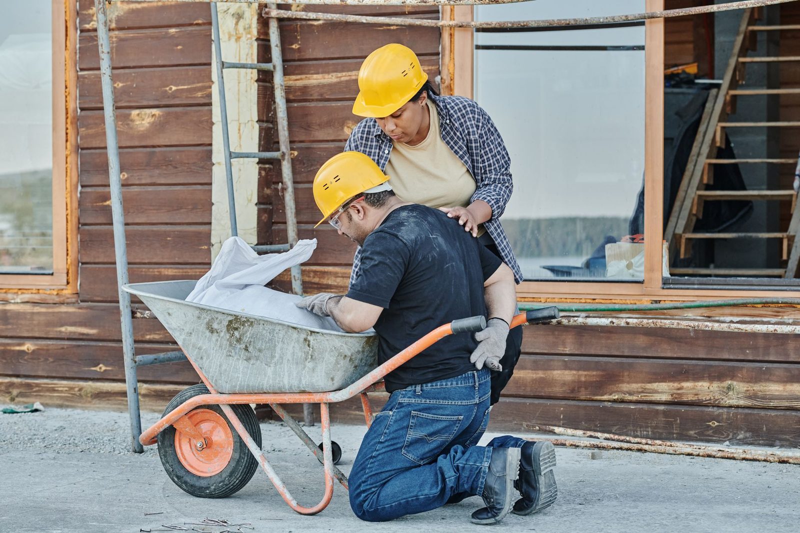 A construction worker kneels beside a wheelbarrow on a job site, appearing to receive help from a colleague.