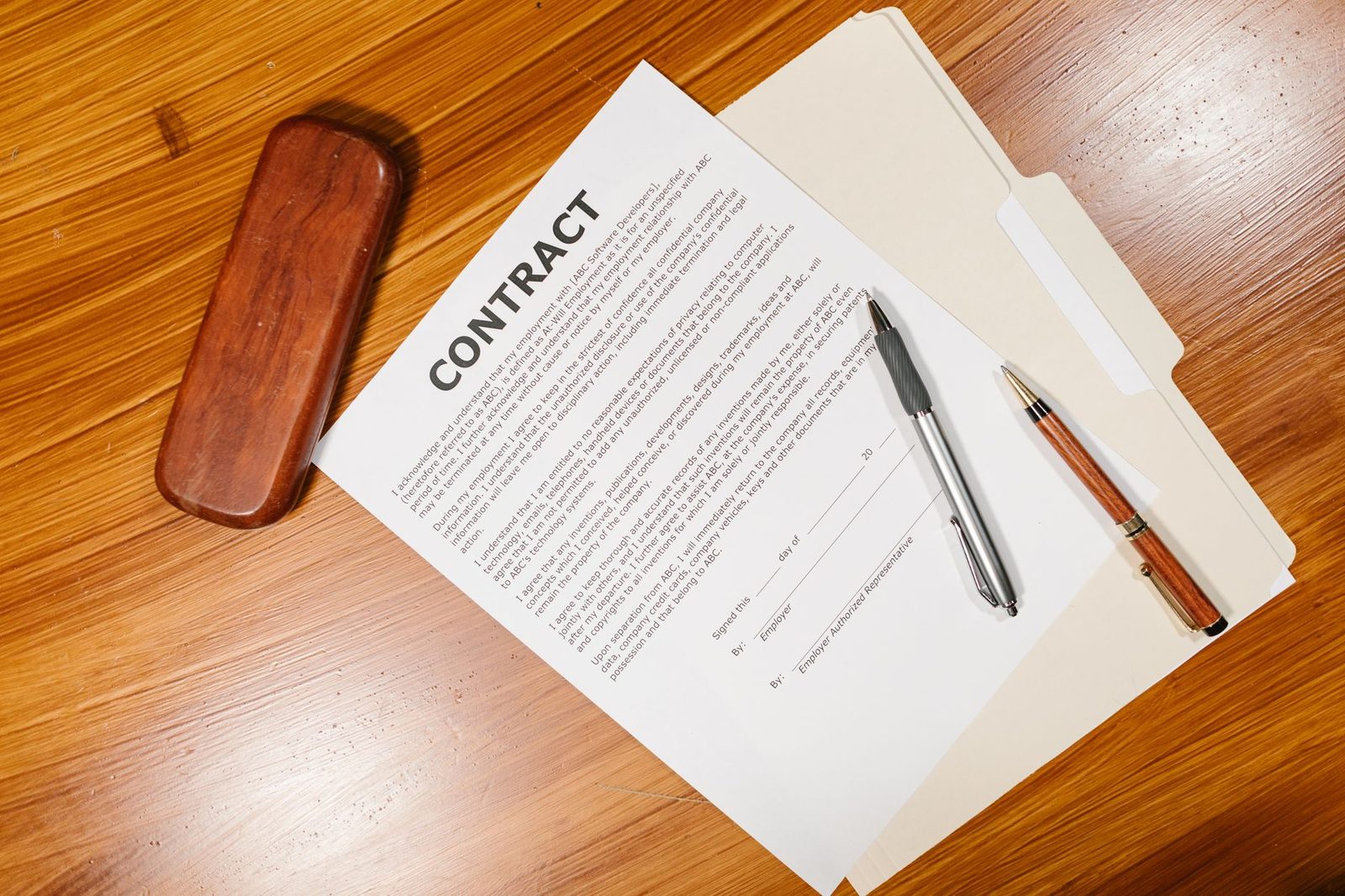 A contract, two pens, and a briefcase sit on a wooden desk.