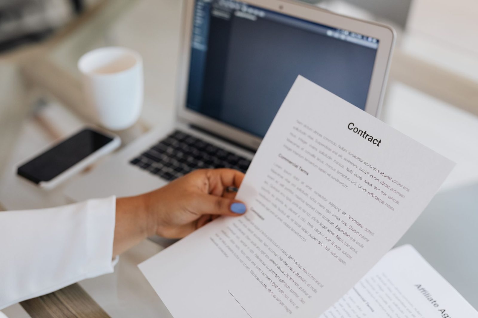 A close-up of a person holding a contract document beside a laptop during a work session.