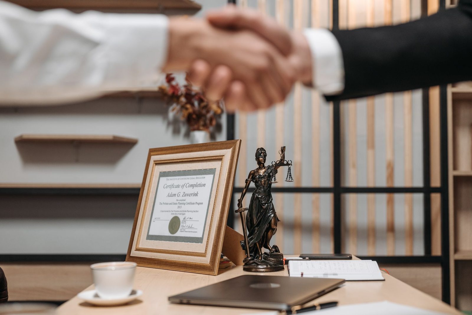 A professional handshake in an office beside a diploma and a Lady Justice statue.