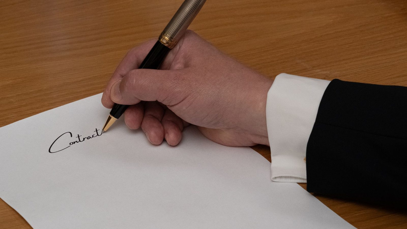 Un profesional firma un contrato de negocios con pluma estilográfica sobre un escritorio de madera.