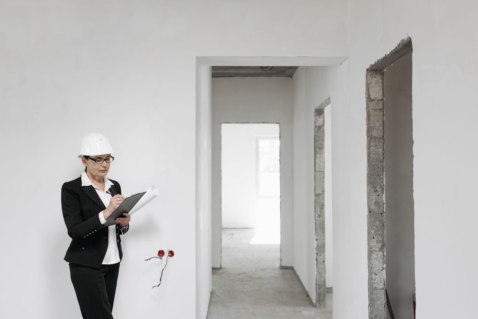A woman in a hard hat and suit inspects an unfinished interior with bare white walls.