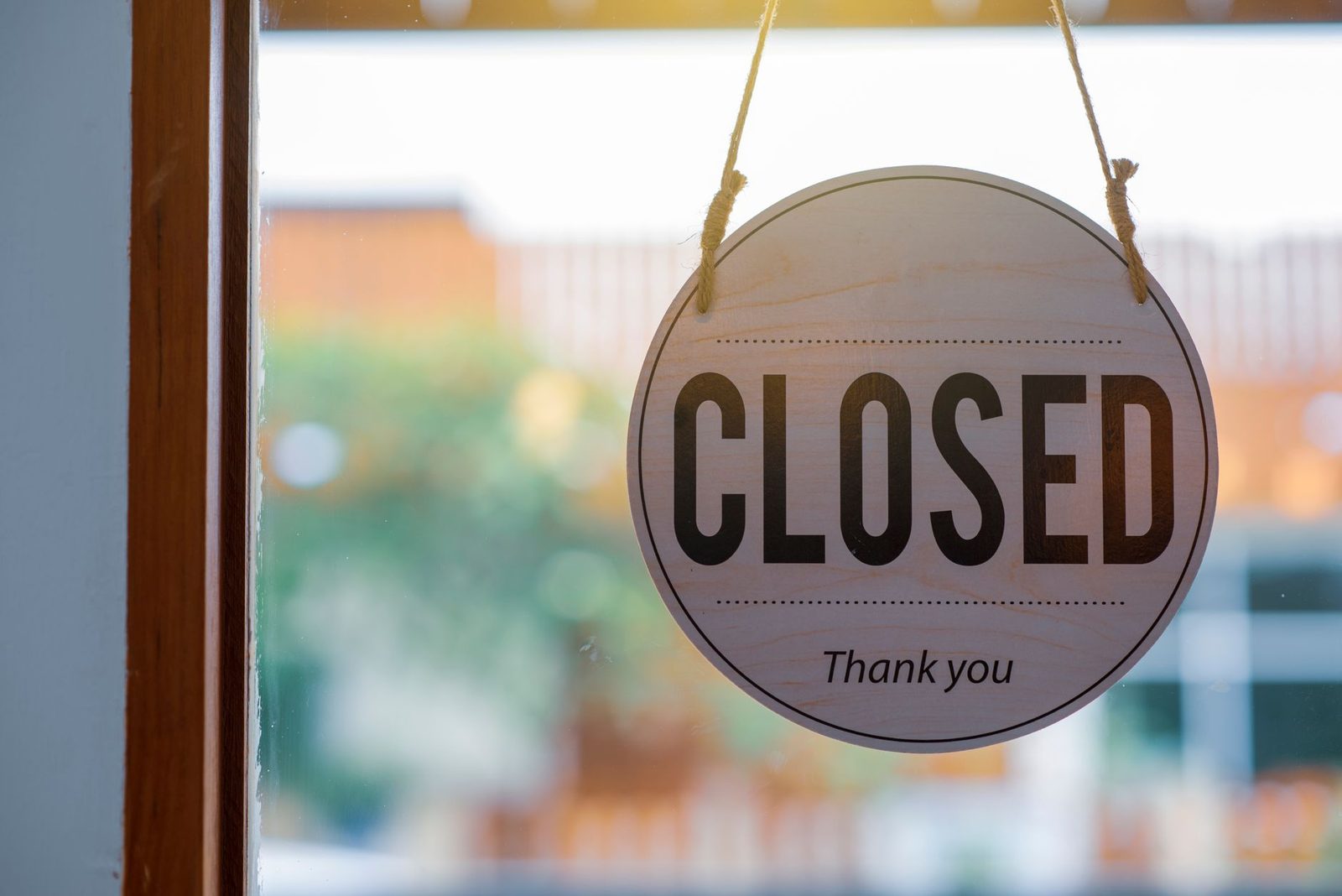 A wooden "Closed" sign hangs in a shop window with a blurred street scene behind it.