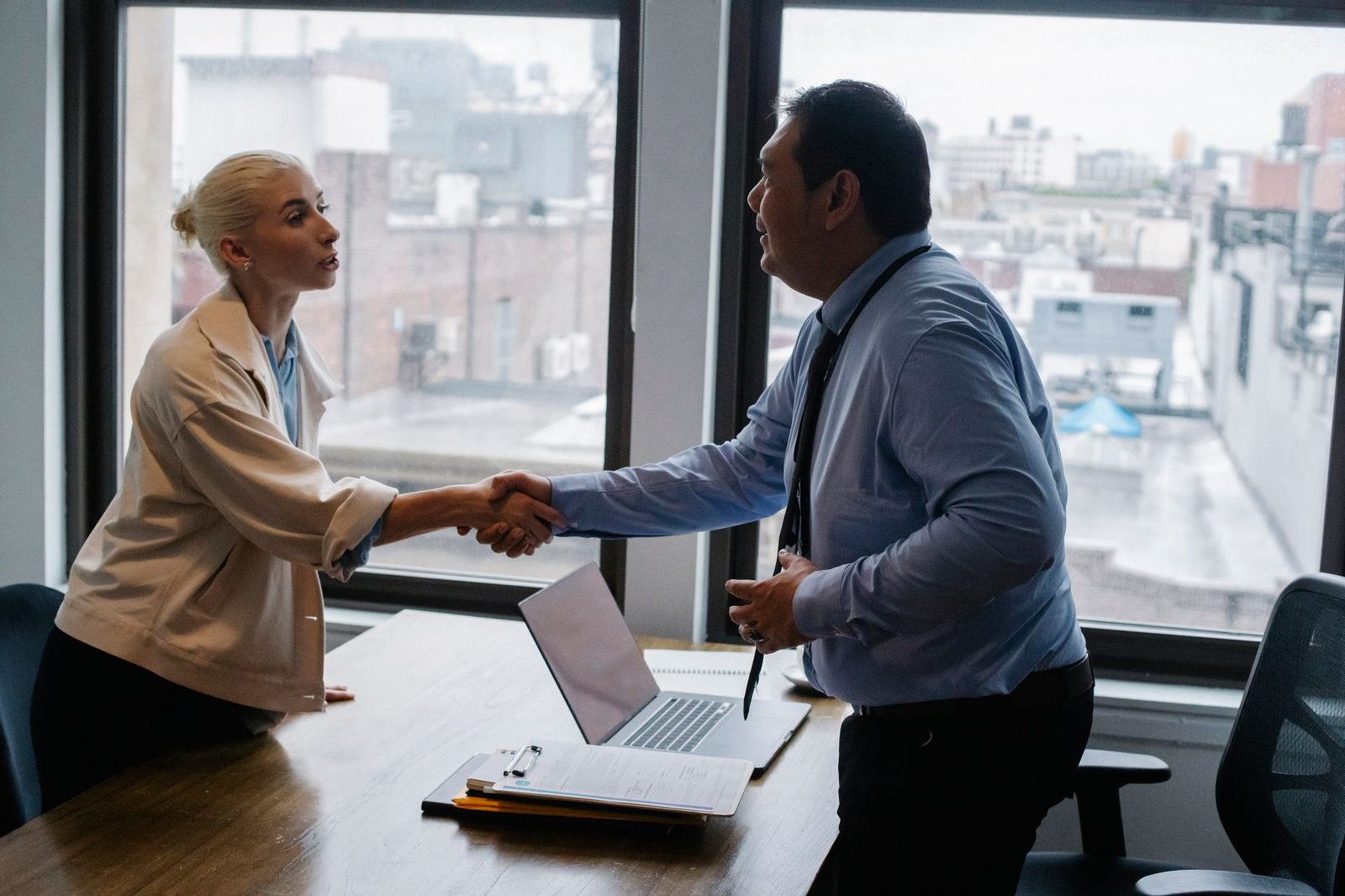 A young woman shakes hands with a smiling employer in a bright office with city views through the windows.