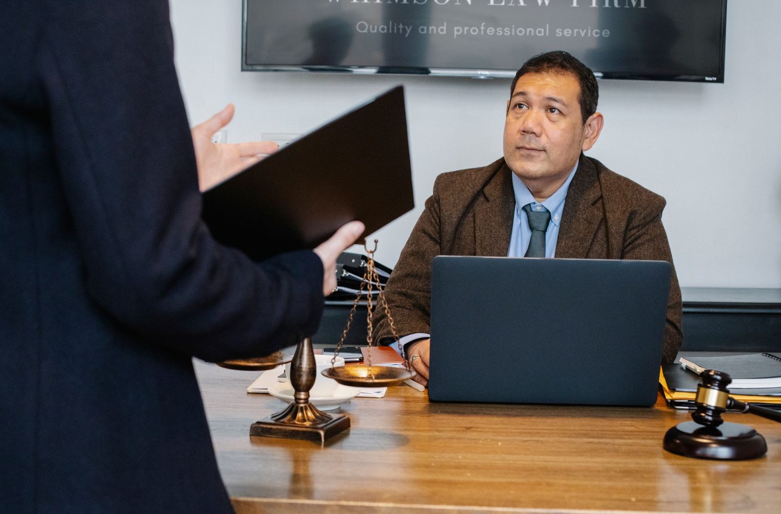 Alt text: A lawyer at a desk with a laptop, gavel, and justice scales as a person shares case details, with the person's face cropped out of view.