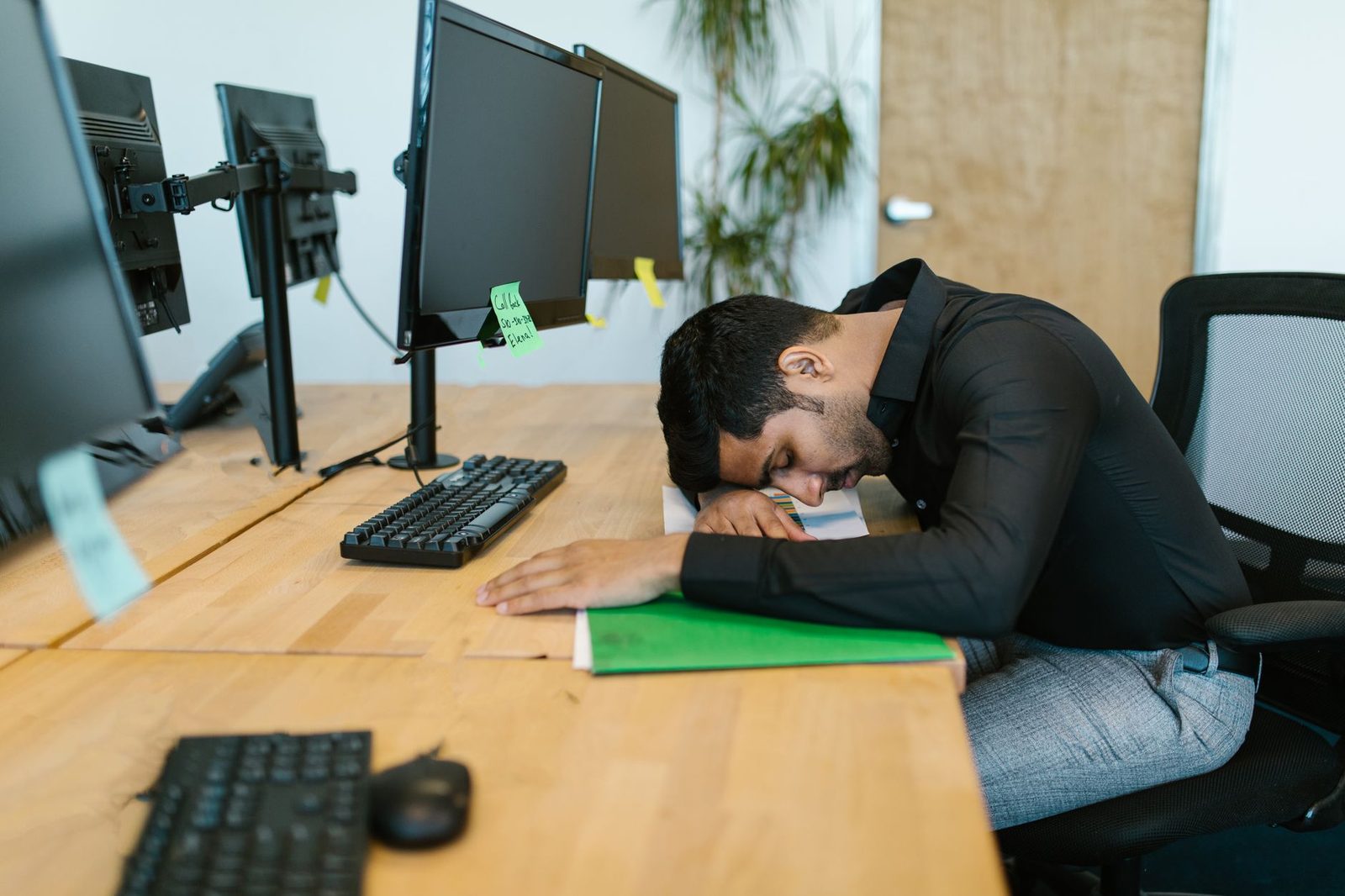 An exhausted office worker rests their head on a desk among multiple computer monitors in a modern workspace.