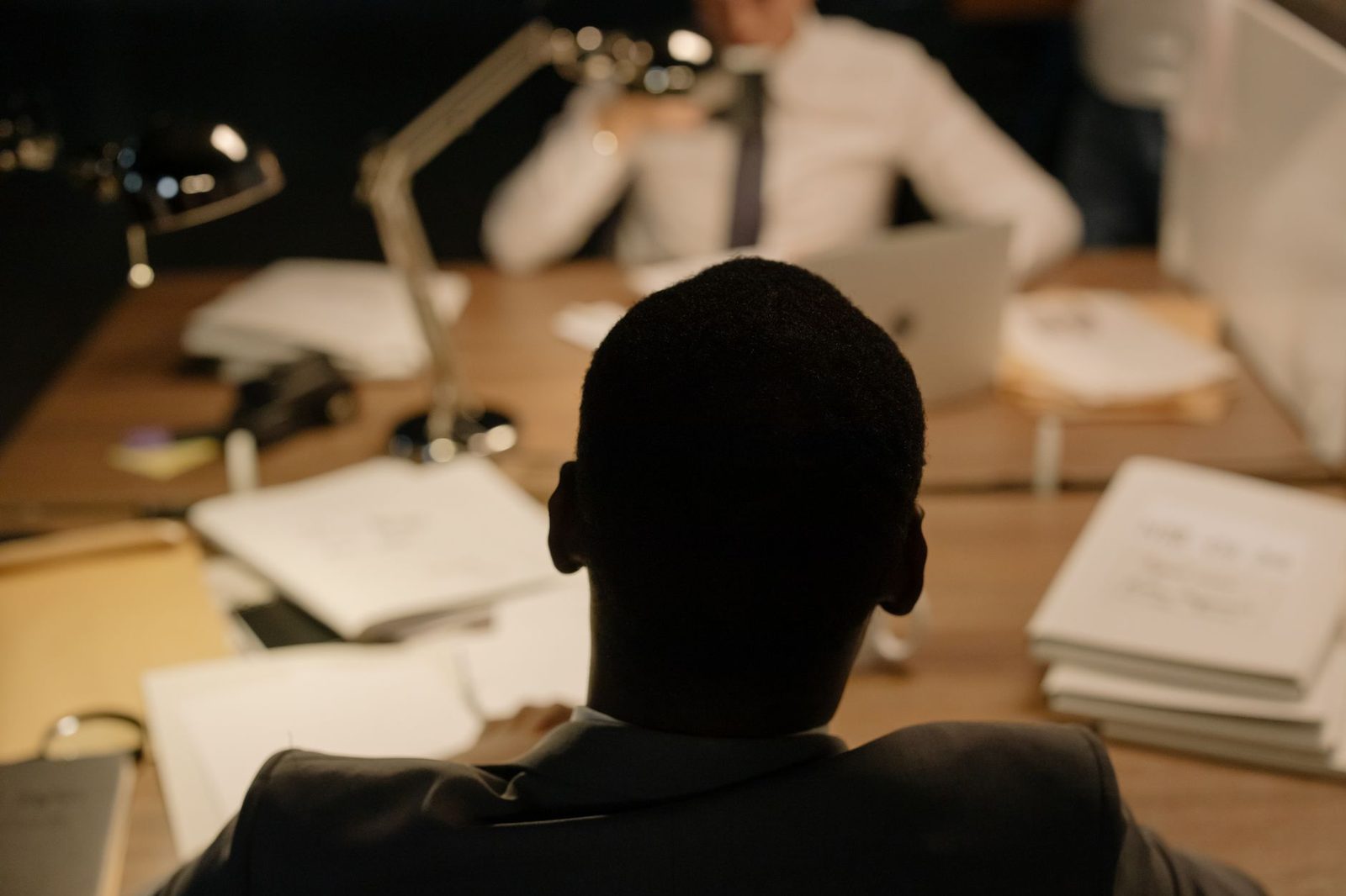 Back view of a professional in a dimly lit office during a business meeting, with documents and laptops on the table.