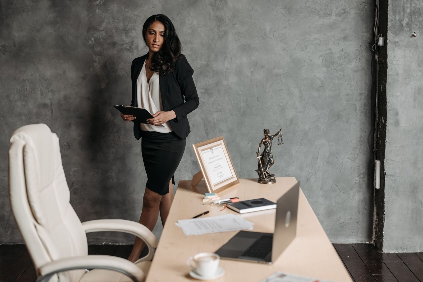 A businesswoman in a formal office reviews legal papers beside a statue of justice on the desk.