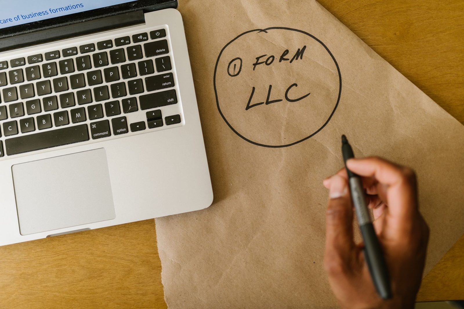 Mano escribiendo "Form LLC" en papel kraft junto a una laptop.