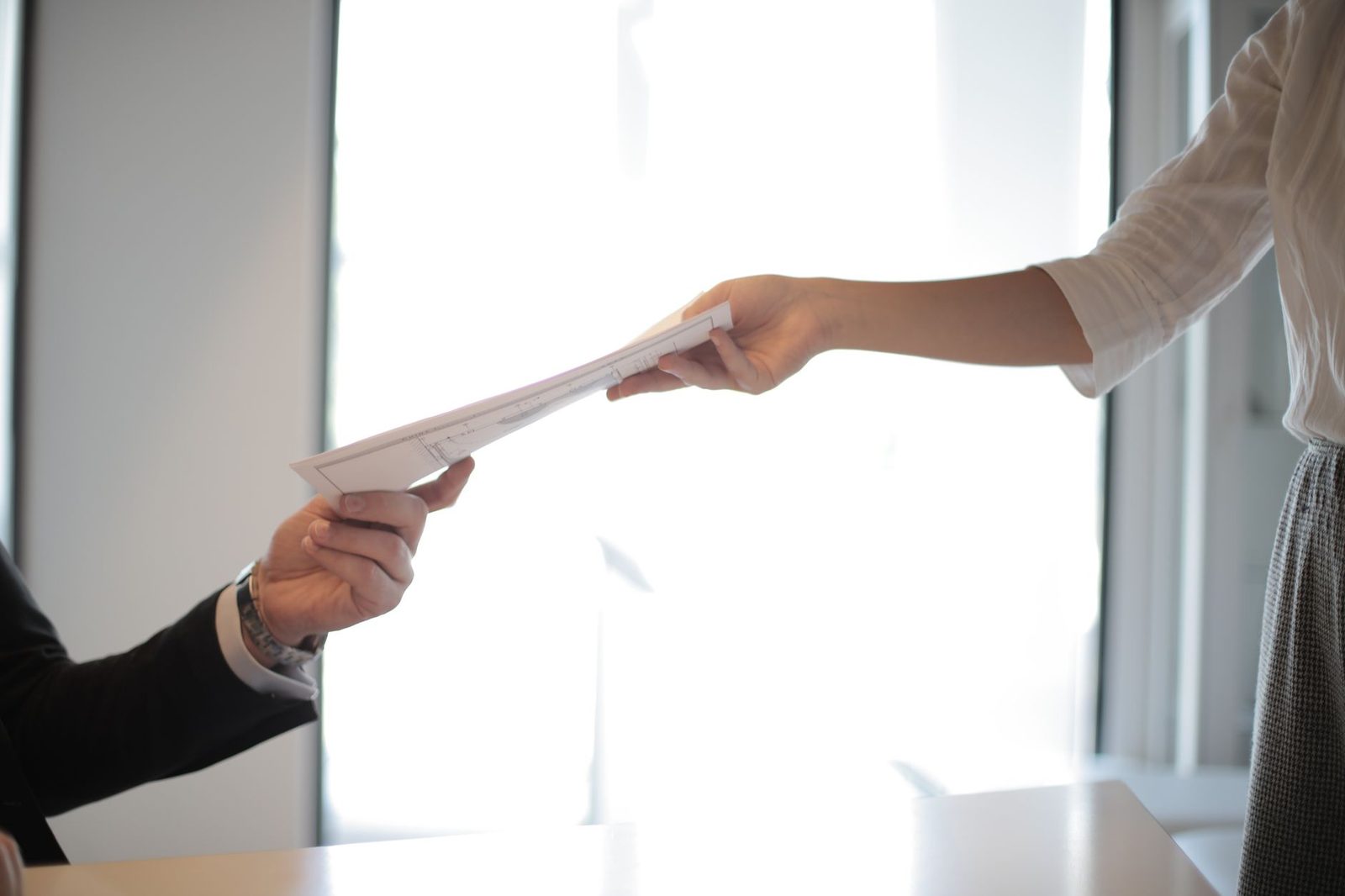 Hands passing paperwork across a desk in an indoor office setting.