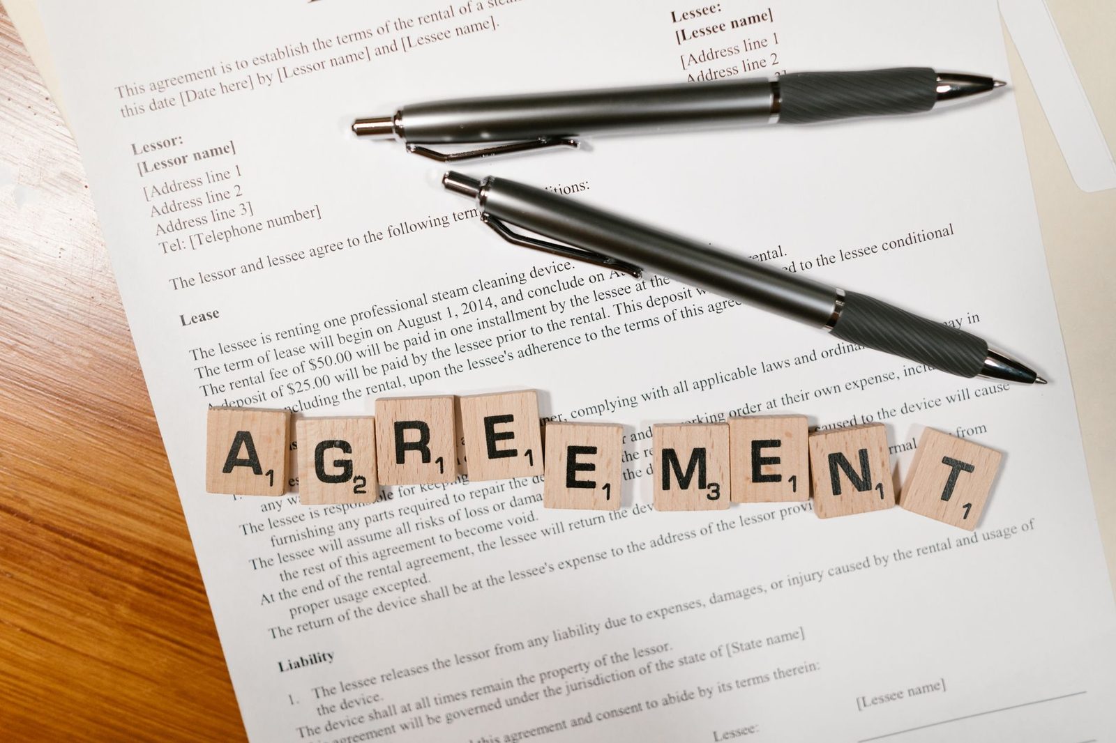 Fichas de Scrabble que deletrean "AGREEMENT" junto a un documento y dos bolígrafos sobre un escritorio.