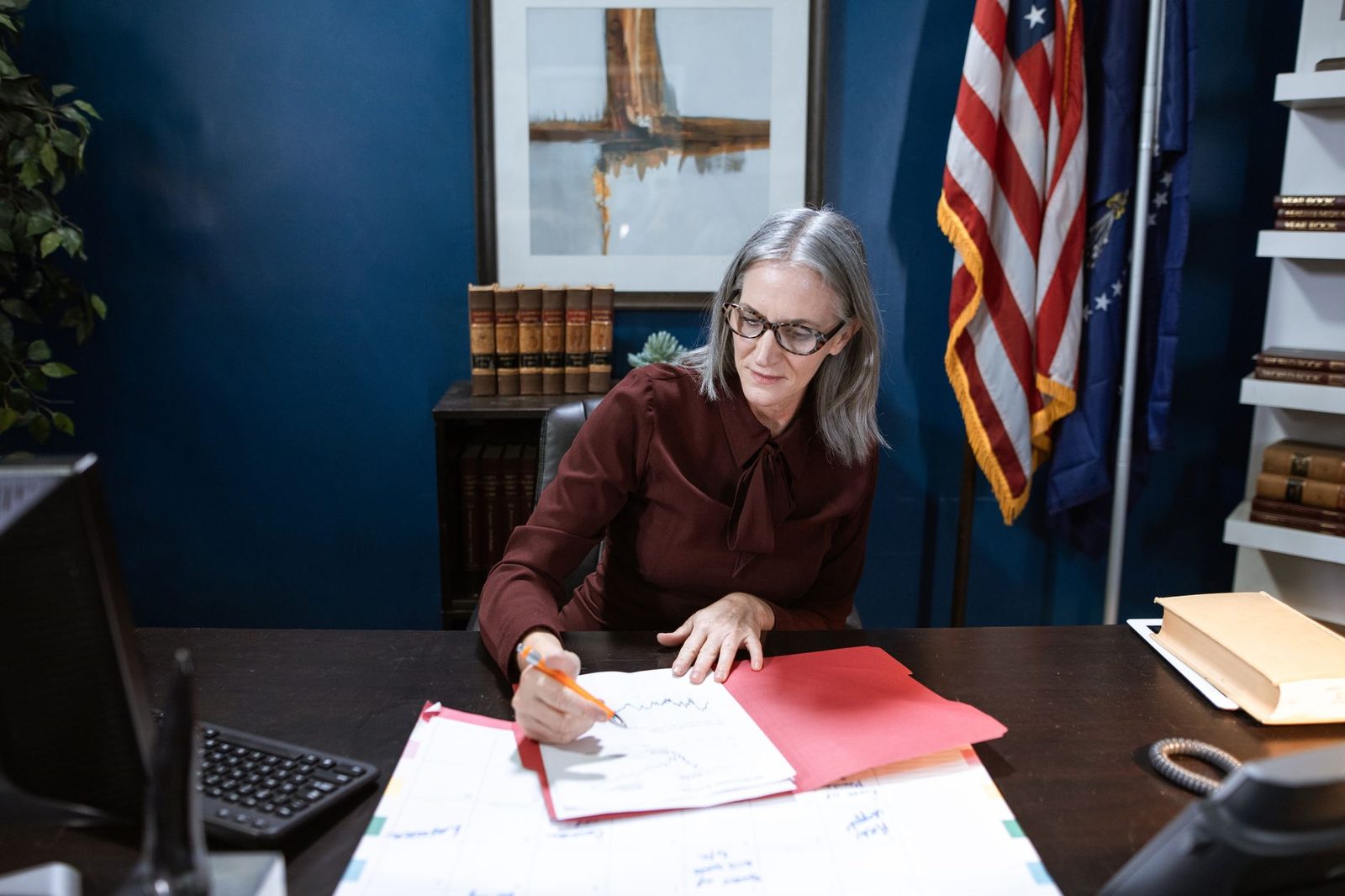 Mujer mayor revisando documentos en un escritorio de oficina, con una bandera de Estados Unidos al fondo.