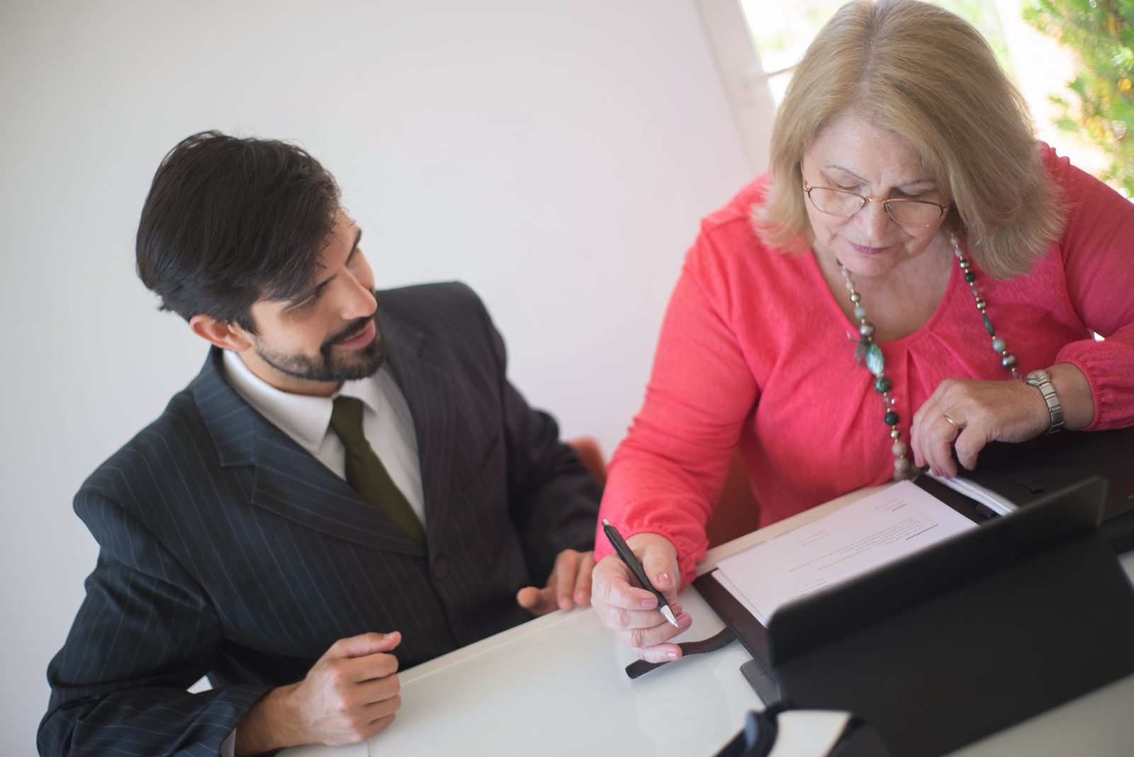 Senior woman signing legal documents with professional assistance at desk.