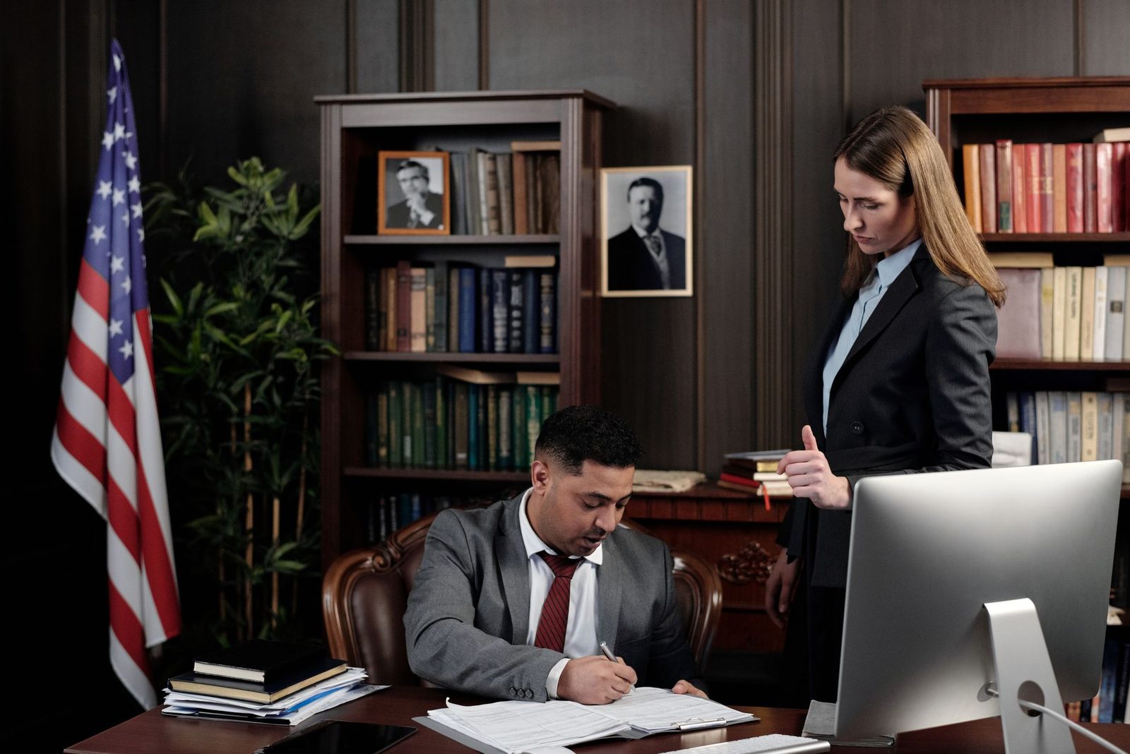 Two business professionals review documents together at a desk in an office with an American flag behind them.