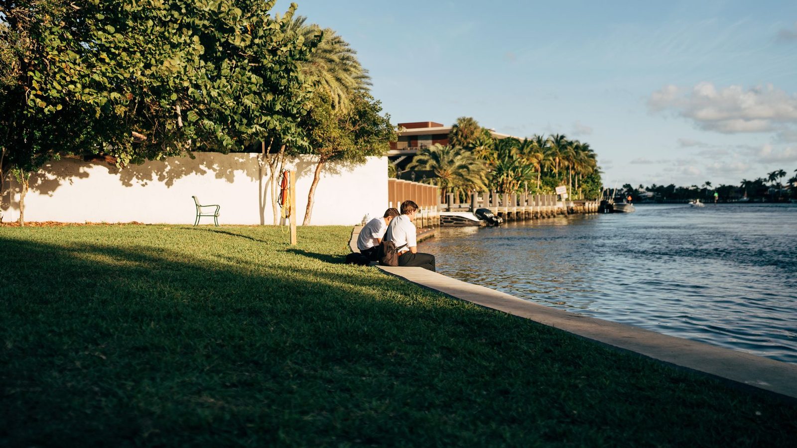 Two people sit by a riverside under palm trees on a sunny day.