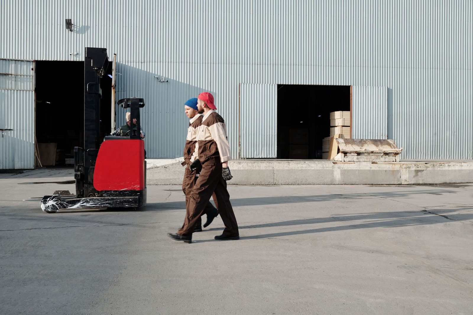 Two uniformed workers walk outside a warehouse past a forklift.