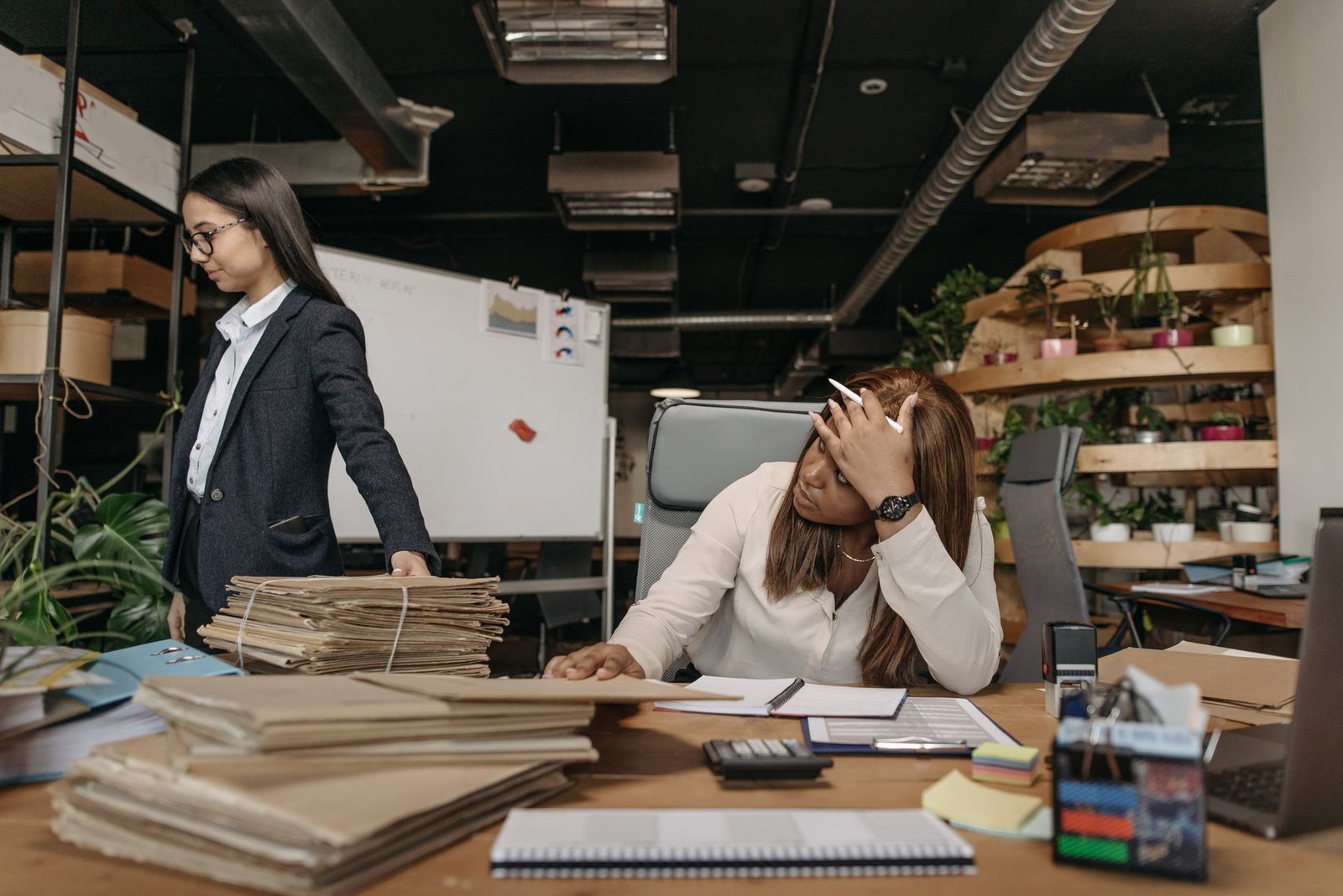 Two women review documents together in a modern office, one seated at a desk and one standing beside her.