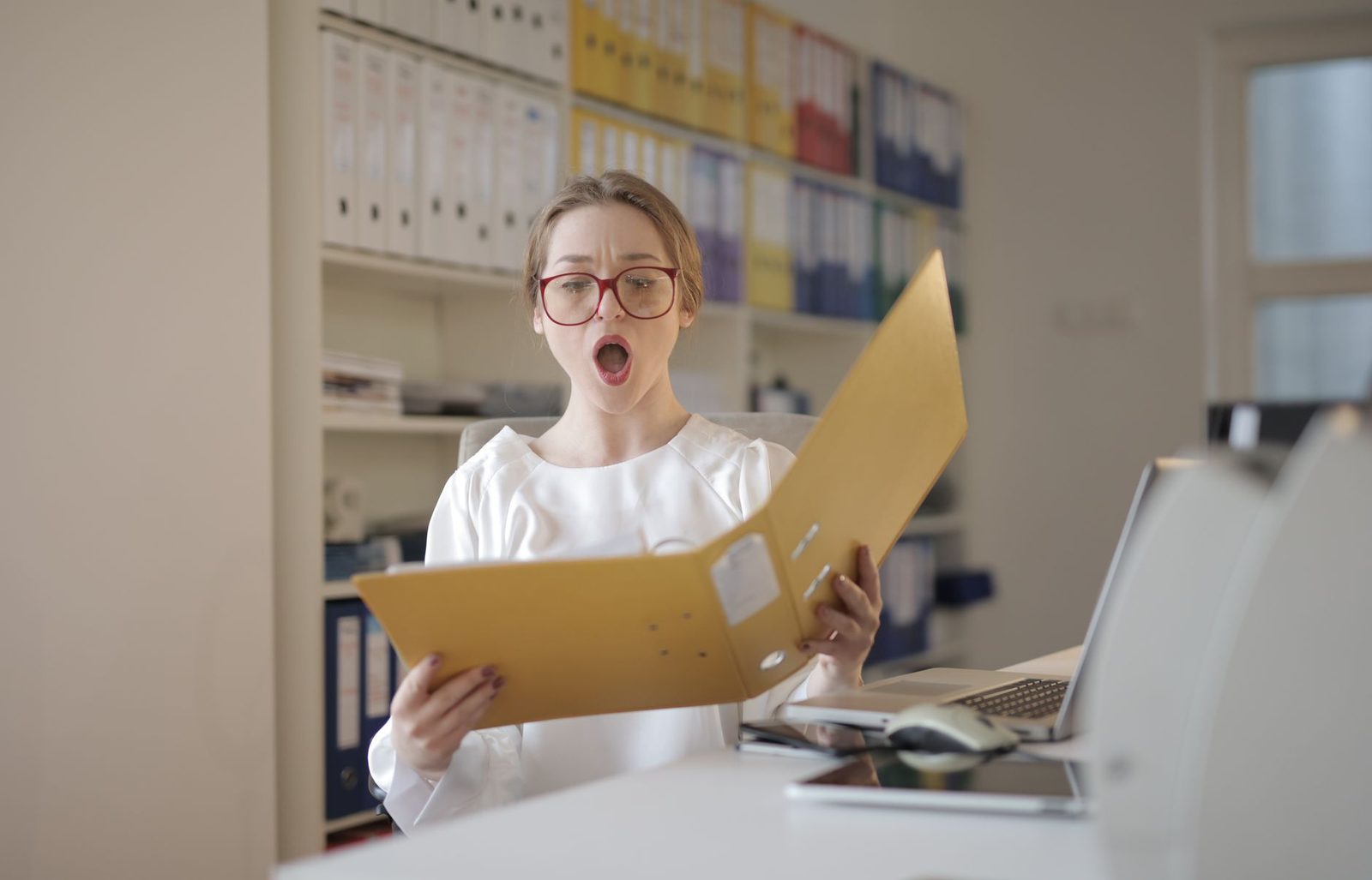 Woman wearing eyeglasses looks surprised while reading a file in an office lined with shelves of records.