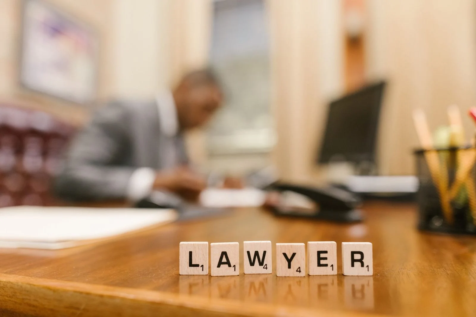 Wooden letter tiles spelling "LAWYER" sit on a desk with a blurred office in the background.